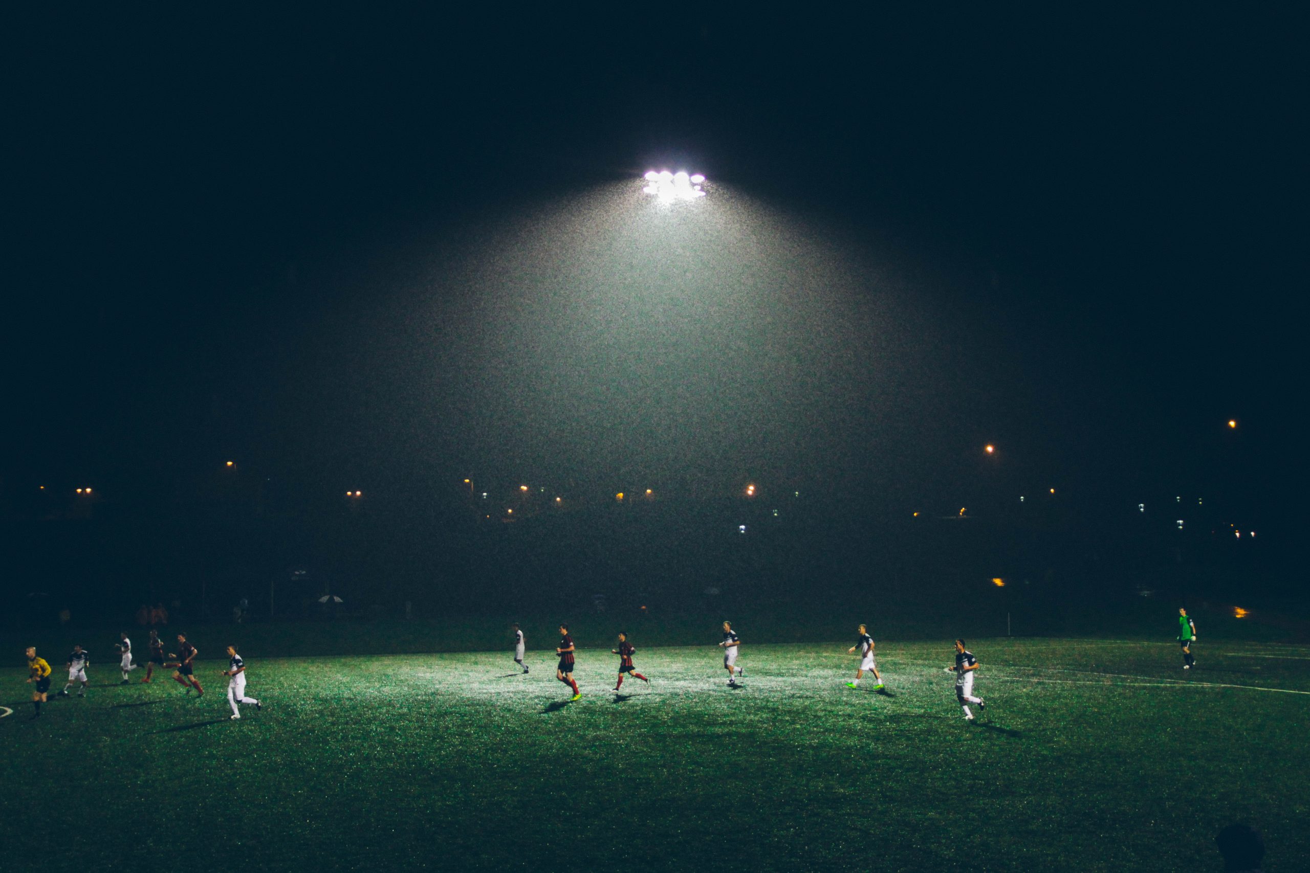 Flood lit football pitch at night with players playing on turf.