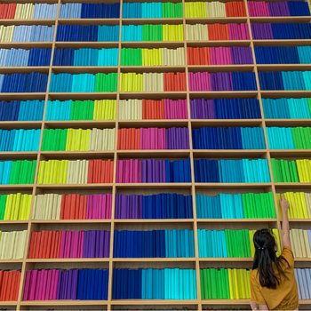 a woman placing a book into a large bookcase with a rainbow of colourful books