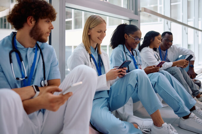 Group of diverse medical and nursing students using their cell phones while relaxing in hallway at university hospital.